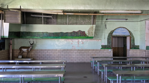 A simple cafeteria with metal tables, a mural of mountains, and a decorative arch on the wall.