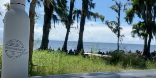 A water bottle sits on a table with a scenic view of trees and a lake in the background under a blue sky.