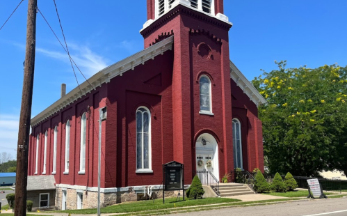 A red brick church with tall windows and a bell tower, surrounded by greenery and a clear blue sky.