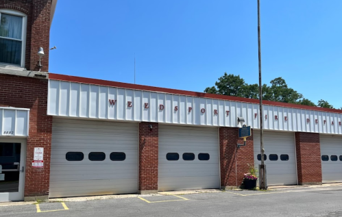 Exterior of a brick building with three garage doors, featuring the sign "Weedsport Fire Department" above. Clear blue sky.