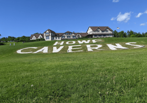 A grassy hillside with large white letters spelling "HOWF CAVERNS" in front of a building under a clear blue sky.