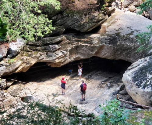 A group of hikers explores a rocky cave surrounded by trees in a natural setting.