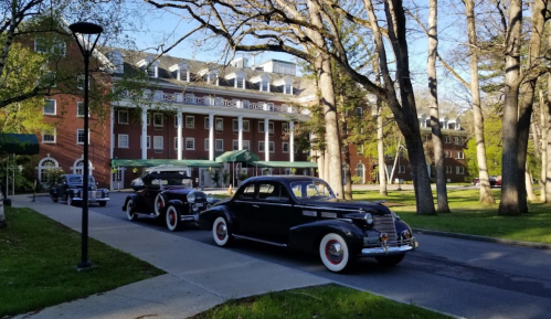 Classic cars drive past a historic brick building surrounded by trees on a sunny day.