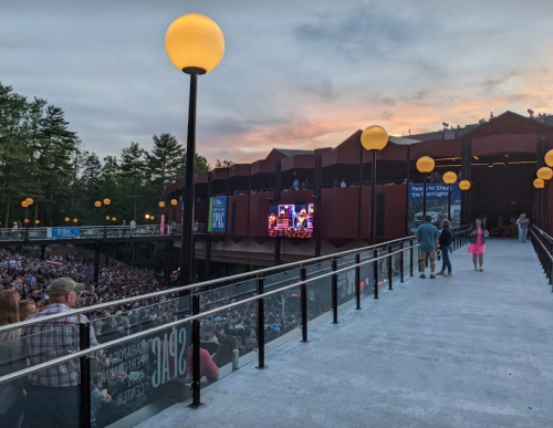 A crowd gathers at an outdoor concert venue during sunset, with bright yellow lights illuminating the walkway.