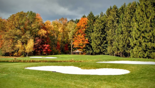A scenic golf course with sand traps, surrounded by vibrant autumn trees and a cloudy sky.
