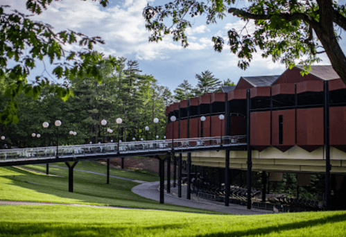 A modern building with a curved walkway, surrounded by green grass and trees, under a partly cloudy sky.