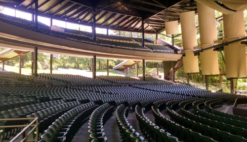 An empty outdoor amphitheater with rows of green seats and a wooded backdrop.