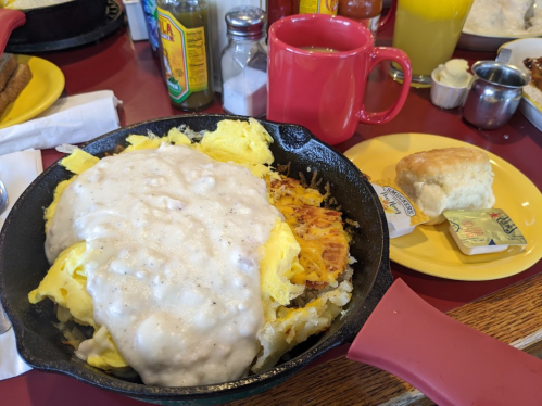 A skillet filled with eggs and gravy over hash browns, alongside a biscuit and a cup of coffee on a red table.