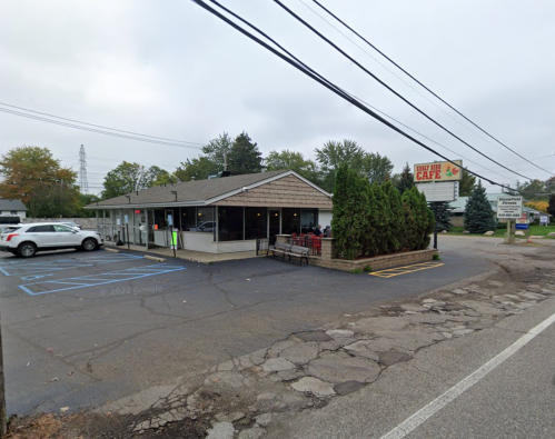 A small café with a sign, surrounded by trees and parked cars, on a cloudy day.
