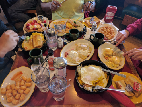 A table filled with various breakfast dishes, including eggs, toast, fruit, and tater tots, with drinks and condiments.