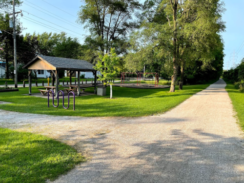 A park scene with a picnic shelter, playground, and a gravel path splitting into two directions, surrounded by greenery.
