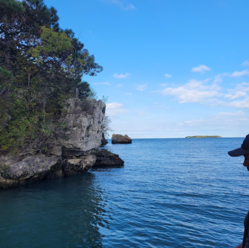 A person stands by the water, gazing at rocky cliffs and a distant island under a clear blue sky.