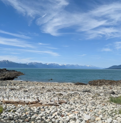 A rocky shoreline with smooth pebbles, blue water, and distant mountains under a clear sky with wispy clouds.