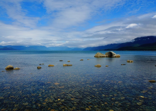 A serene lake scene with clear water, scattered rocks, and mountains under a partly cloudy sky.