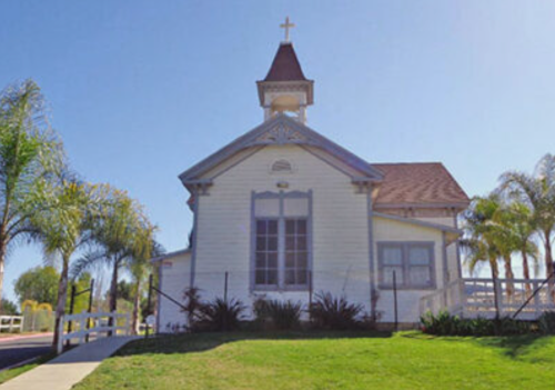 A small white church with a steeple and cross, surrounded by palm trees and green grass under a clear blue sky.
