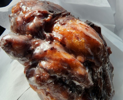 A close-up of a glazed, caramelized apple fritter on white paper, showcasing its shiny, textured surface.