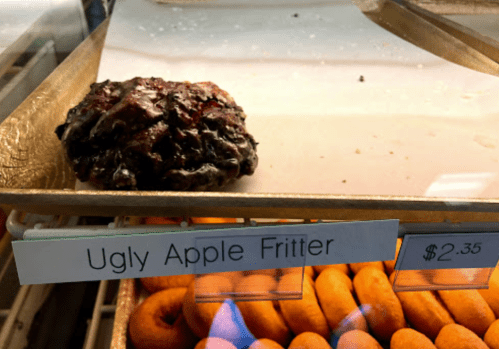A close-up of an "Ugly Apple Fritter" on a display case, with other donuts in the background. Price: $2.35.