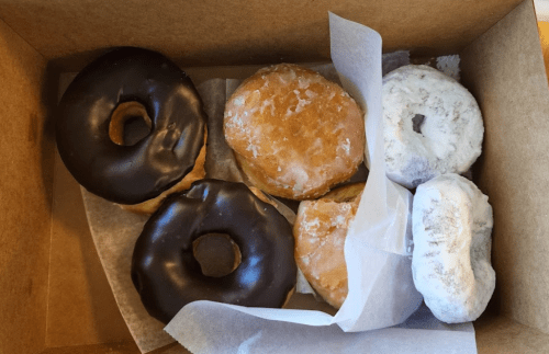 A box of assorted donuts, including chocolate-glazed, glazed, and powdered varieties, arranged on parchment paper.