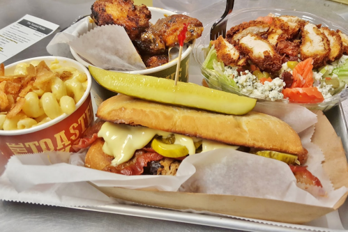 A tray with a sandwich topped with pickles, mac and cheese, chicken wings, and a salad with tomatoes and blue cheese.