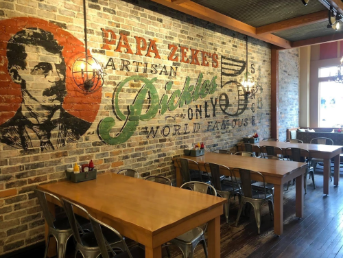 Interior of a restaurant featuring a brick wall mural of a man and the text "Papa Zeke's Artisan Pickles." Tables are set.
