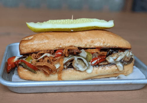 A loaded sandwich with grilled vegetables, melted cheese, and crispy onions, topped with a pickle slice.