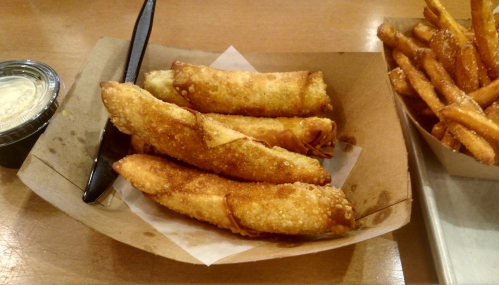 Four crispy spring rolls in a paper tray, accompanied by a small container of dipping sauce and a side of sweet potato fries.
