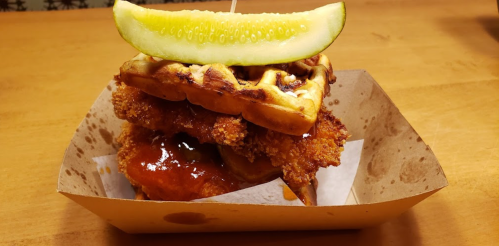 A stacked sandwich featuring crispy fried chicken and waffles, topped with a pickle slice and drizzled with sauce.