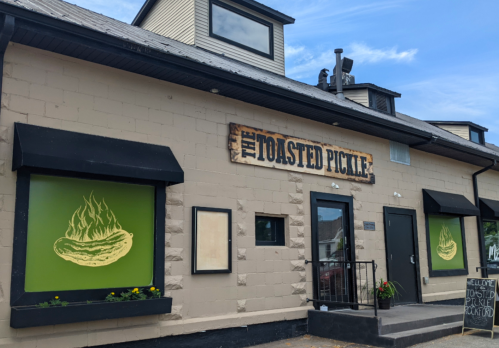 Exterior of "The Toasted Pickle" restaurant featuring a sign and green windows with pickle illustrations.