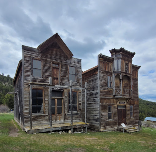 Two weathered wooden buildings from a ghost town, featuring large windows and a rustic facade, set against a cloudy sky.