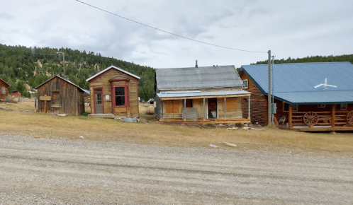 Abandoned buildings in a rural area, with a gravel road and mountains in the background under a cloudy sky.
