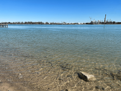 A calm river scene with clear water, a rocky shore, and industrial structures in the background under a blue sky.