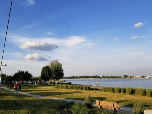 A scenic riverside park with green grass, trees, and people walking along the path under a blue sky with clouds.