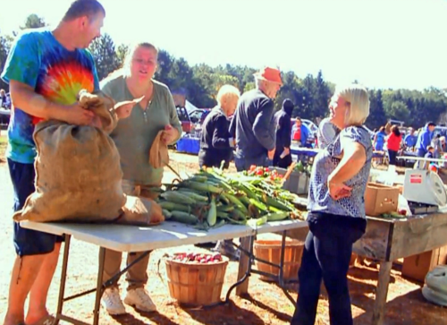 A bustling farmers market scene with people interacting and various fresh vegetables displayed on tables.