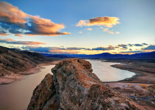 A rocky outcrop overlooks a winding river at sunset, with colorful clouds and mountains in the background.