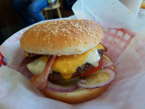 A close-up of a cheeseburger with bacon, lettuce, tomato, pickles, and onions on a sesame seed bun in a basket.