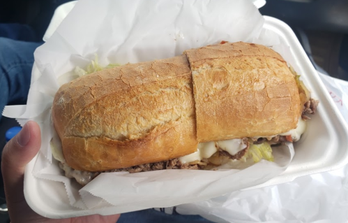 A hand holding a large sandwich in a white container, filled with meat, lettuce, and mayonnaise, on a crinkled paper.
