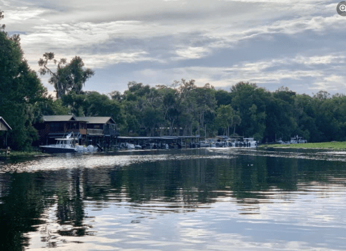 A serene river scene with wooden houses and boats, surrounded by lush greenery under a cloudy sky.