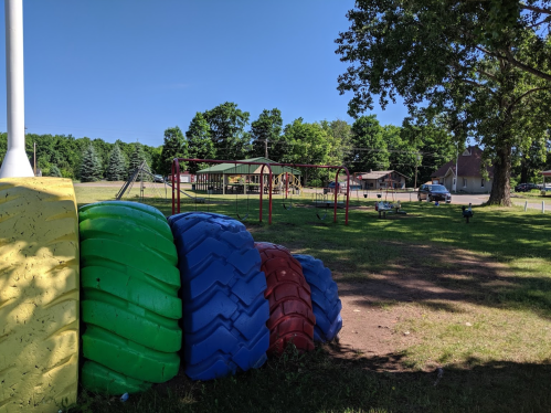 Colorful tire structures in a grassy playground with swings and a pavilion in the background under a clear blue sky.