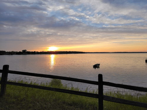 A serene sunset over a calm lake, with a small boat and a wooden fence in the foreground.