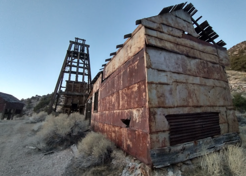 Rusty, abandoned mining structures with a wooden tower and a weathered building, surrounded by dry vegetation.