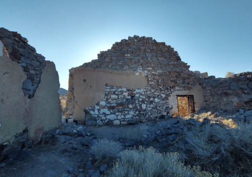 Ruins of a stone building with a partially collapsed roof, surrounded by rocky terrain and sparse vegetation.