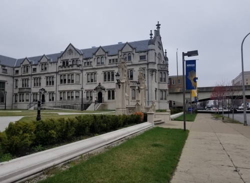 Historic building with ornate architecture, surrounded by greenery and a sidewalk, under a cloudy sky.