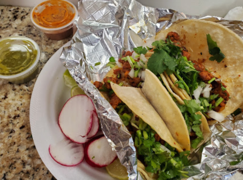 A plate of tacos with cilantro, onions, and radishes, accompanied by two sauces in containers.