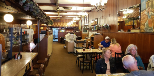 A cozy restaurant interior with wooden walls, tables, and patrons dining. Light fixtures and decor add a warm ambiance.