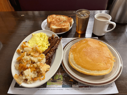 A breakfast plate with scrambled eggs, bacon, hash browns, pancakes, toast, and coffee on a table.