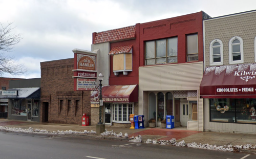 A small town street featuring a restaurant, shops, and snow along the sidewalk under a cloudy sky.
