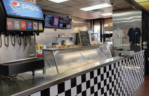 A fast-food counter with a Pepsi dispenser, menu board, and food display in a checkered-tile setting.
