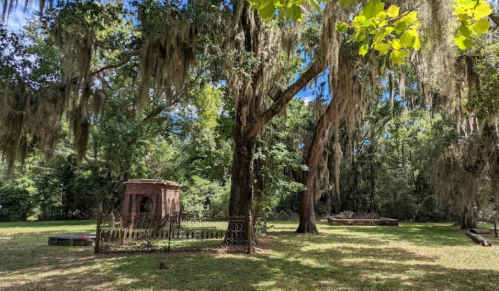 A serene cemetery scene with moss-covered trees and a small, ornate structure surrounded by a wrought-iron fence.