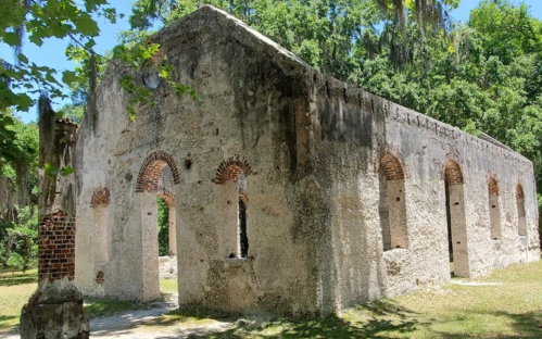 Ruins of an old stone building surrounded by trees and greenery under a clear blue sky.