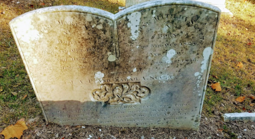 Weathered gravestone with inscriptions, partially obscured, set in a grassy cemetery.